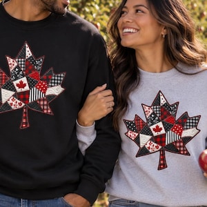 May include: A black crewneck sweatshirt and a light gray crewneck sweatshirt, each featuring a large, central maple leaf graphic composed of a patchwork of red, black, and white plaid, gingham, and solid fabric patterns with stitched detailing.