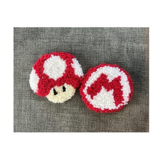 Puede incluir: Dos posavasos hechos a mano con forma de hongo rojo, blanco y beige. Un posavasos presenta un diseño de hongo con ojos negros, y el otro tiene un diseño de M rojo y blanco. Los posavasos están hechos de un material texturizado.