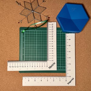 Peut inclure: Un ensemble d'outils et de fournitures d'artisanat, comprenant un tapis de découpe vert, des règles avec des mesures métriques et impériales, et un objet hexagonal bleu avec "20°" imprimé. Un ornement décoratif en forme de flocon de neige est également présent.