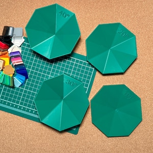 May include: Four green, octagonal tools marked with angles (10°, 20°, 30°, 40°). A green cutting mat with cm markings is visible, alongside a colour swatch of various shades. The tools are likely for crafting.