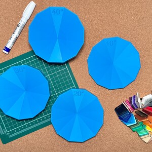 Könnte beinhalten: Vier blaue Winkelmesser mit den Markierungen 20°, 30°, 40° und 70° liegen auf einer Korkplatte. Ein weißer Marker und eine grüne Schneidematte sind ebenfalls vorhanden. Ein Farbmusterfächer befindet sich unten rechts.
