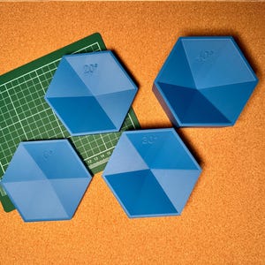 Puede incluir: Cuatro objetos geométricos azules con superficies anguladas, cada uno marcado con una medida en grados (20°, 30°, 40° y 50°). Una esterilla de corte verde está en el fondo, y los objetos están sobre una superficie marrón.