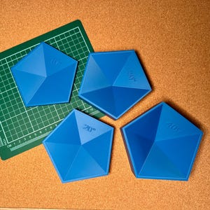 以下が含まれることがあります： 角度の付いた側面を持つ、青色の幾何学的な五角形のツールが4つ。それぞれに角度の測定値がマークされています。1つはグリッドパターンの緑色のカッティングマットの上に、他は茶色のコルクボードの表面にあります。これらのツールは、クラフトやデザイン用と思われます。