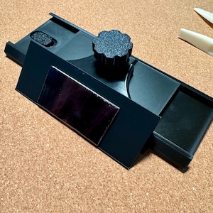 May include: A black 3D printed tool featuring a rectangular base with a sliding mechanism, a central circular knob with a textured grip, and a smooth, reflective rectangular plate attached to the front face. The device has a matte finish.
