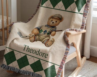 May include: A beige throw blanket with a green diamond pattern and fringe, draped over a wooden rocking chair. The blanket features a teddy bear wearing golf attire and holding a golf club. The name "Theodore" is printed on the blanket.