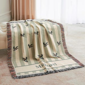 May include: A striped throw blanket in cream and sage green, featuring embroidered duck motifs. The blanket has a fringed edge and the name "Michael" embroidered on a band. The blanket is draped over a cream-colored ottoman.
