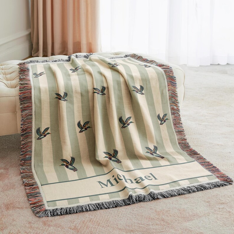 May include: A striped throw blanket in cream and sage green, featuring embroidered duck motifs. The blanket has a fringed edge and the name "Michael" embroidered on a band. The blanket is draped over a cream-colored ottoman.