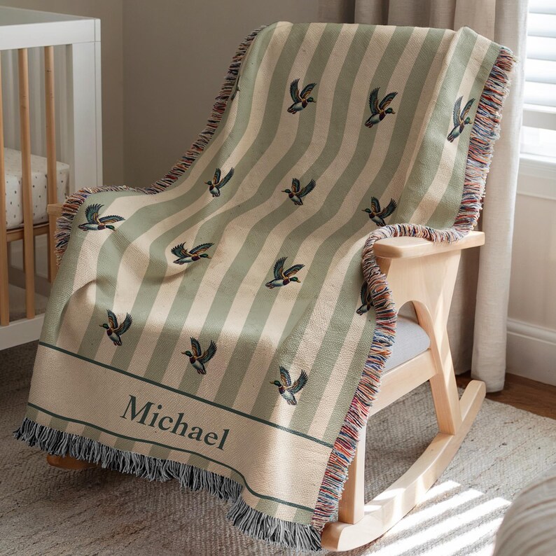 May include: A striped, sage green and cream-colored throw blanket with a duck pattern, draped over a wooden rocking chair. The blanket has a personalized name, "Michael", and fringed edges.