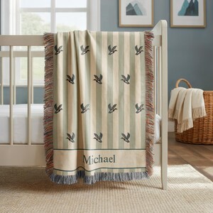 May include: A beige and green striped baby blanket with a duck pattern and the name "Michael" embroidered on it. The blanket has a fringed edge and is draped over a crib. The blanket is made of a soft material.