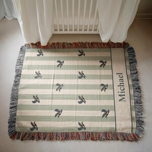May include: A rectangular woven blanket with a striped pattern in cream and light green. The blanket features a repeating bird design and a personalized name, "Michael," in a vertical column. The edges are fringed with a mix of colors.