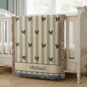 May include: A personalized baby blanket with a duck pattern. The blanket features vertical stripes in cream and sage green, with embroidered ducks. The name "Michael" is displayed in a banner at the bottom. The blanket is draped over a white crib.