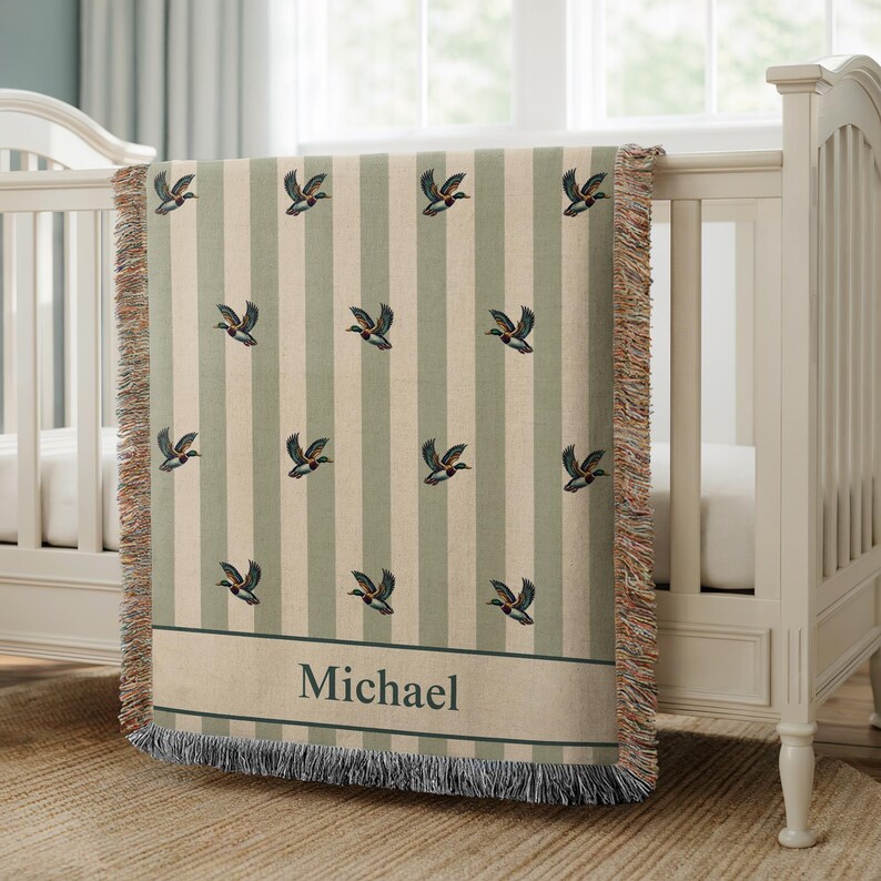May include: A personalized baby blanket with a duck pattern. The blanket features vertical stripes in cream and sage green, with embroidered ducks. The name "Michael" is displayed in a banner at the bottom. The blanket is draped over a white crib.