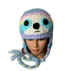 May include: A crocheted hat with a sloth face design. The hat is made with a rainbow of colors, including pink, blue, yellow, and white. The hat has ear flaps and a long braided tie.