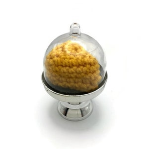 May include: A small crocheted yellow hat displayed under a clear glass dome on a silver stand.