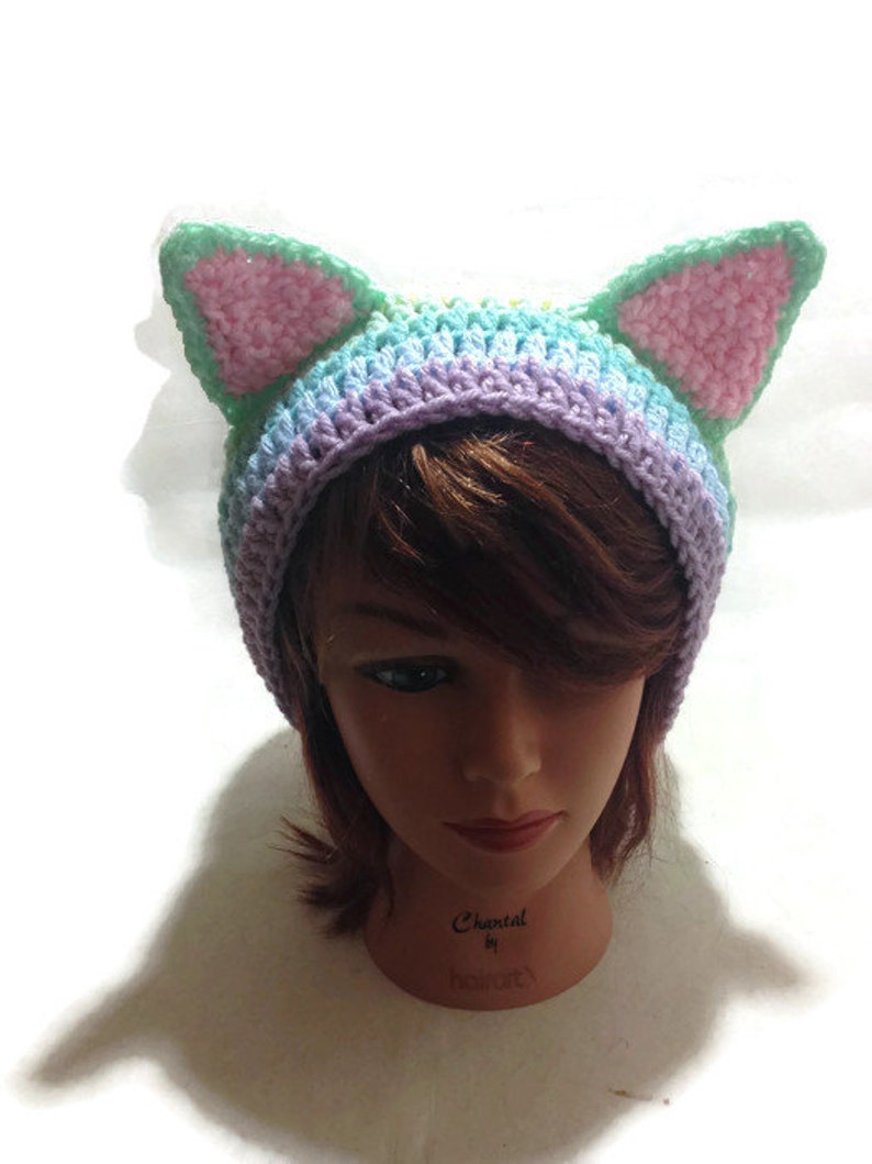 May include: A crocheted hat with cat ears in shades of purple, blue, green, and pink. The hat is on a mannequin head.