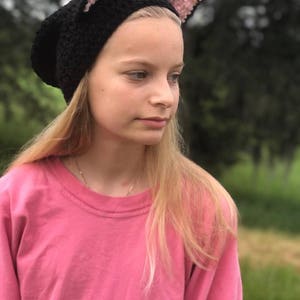May include: A black crocheted beanie with pink cat ears. The beanie is being worn by a person with long blonde hair and a pink shirt.