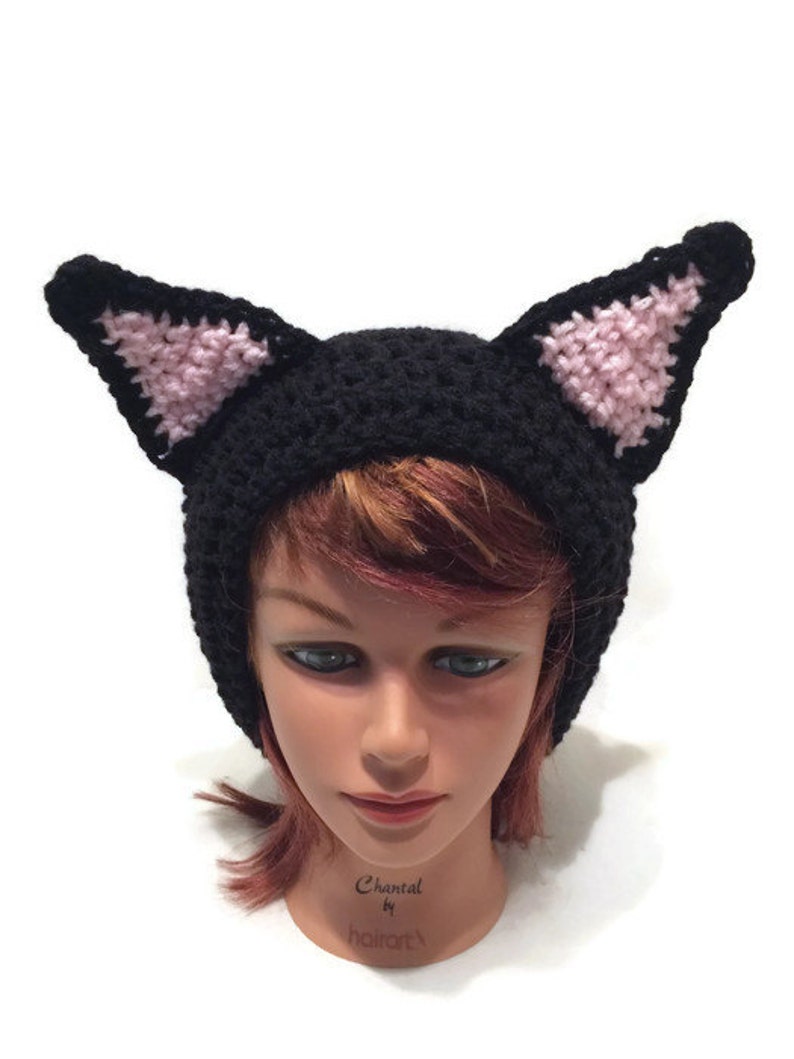 May include: Black crocheted cat hat with pink ears. The hat is on a mannequin head with red hair.
