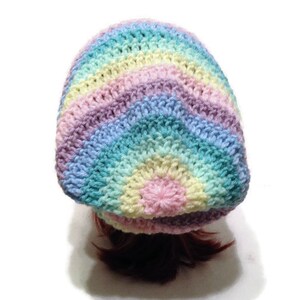 May include: A rainbow crochet beanie with a long ponytail coming out of the top. The beanie is made with pastel colors of pink, blue, green, and yellow.