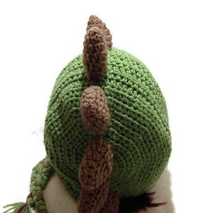 May include: Green and brown crocheted dinosaur hat with a long tail. The hat has a pointed top and a textured surface.