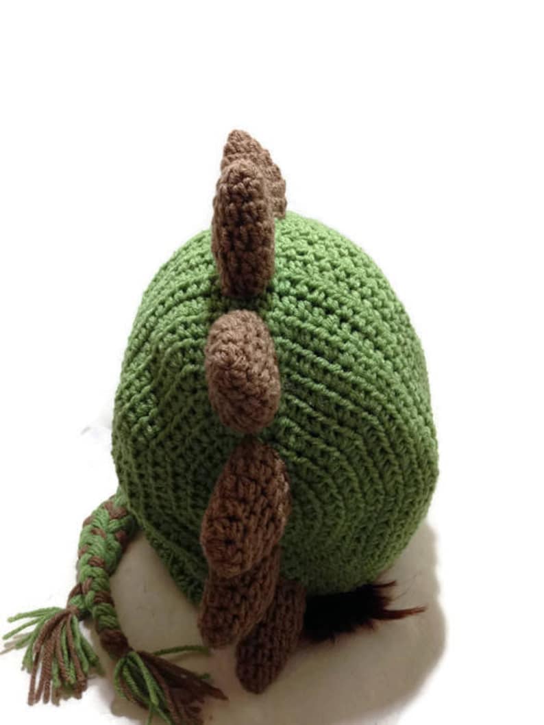 May include: Green and brown crocheted dinosaur hat with a long tail. The hat has a pointed top and a textured surface.
