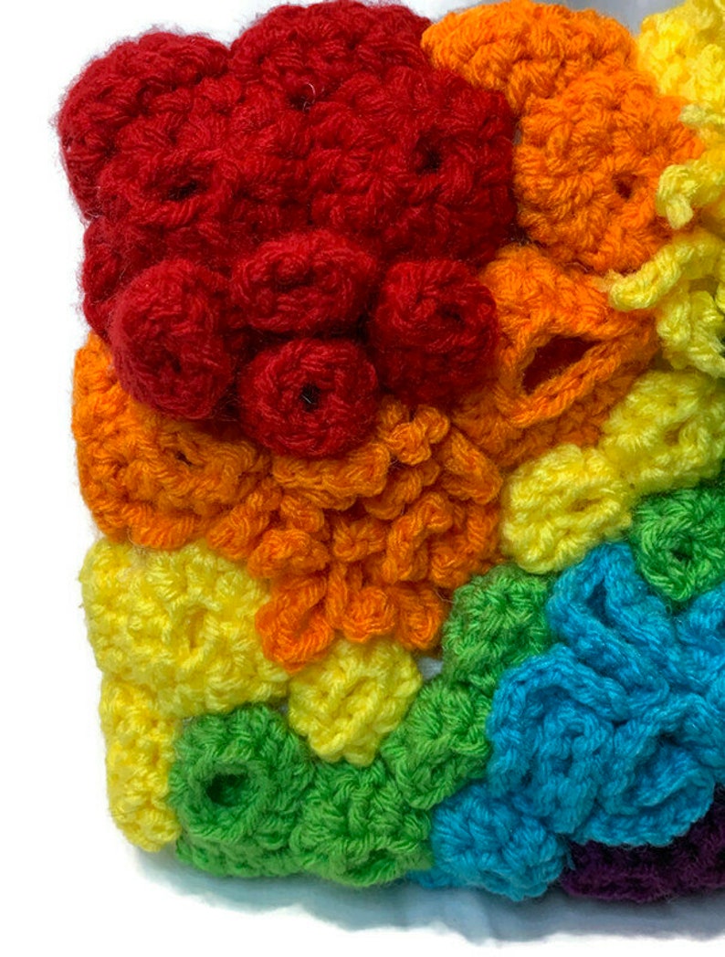May include: A colourful crocheted square with a rainbow pattern. The square is made of different coloured yarn, including red, orange, yellow, green, blue, and purple.