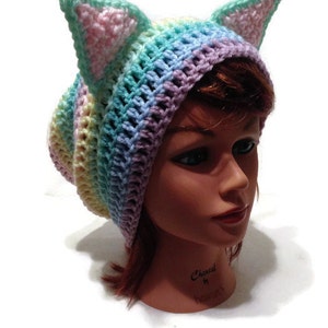 May include: A pastel rainbow colored crochet beanie with cat ears. The beanie is made with a variety of colors, including pink, blue, green, and yellow.