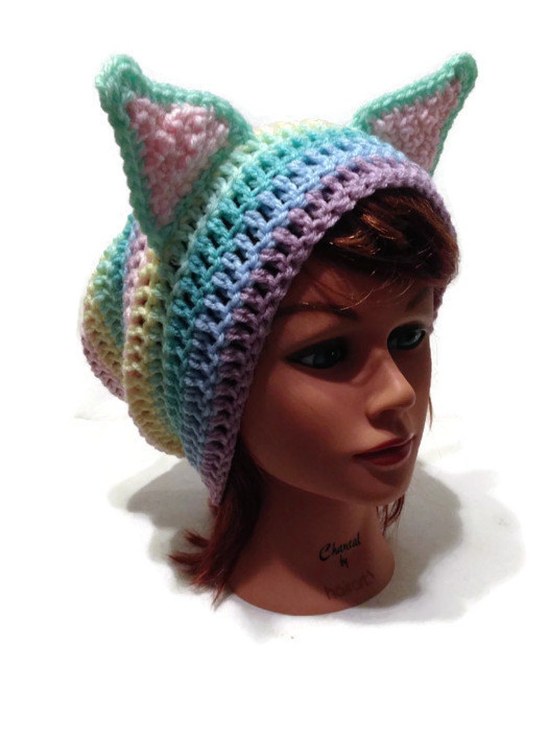 May include: A pastel rainbow colored crochet beanie with cat ears. The beanie is made with a variety of colors, including pink, blue, green, and yellow.
