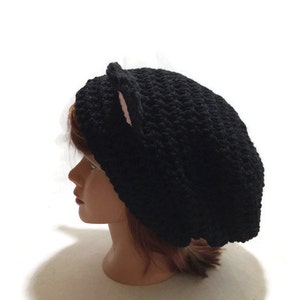 May include: Black crocheted beanie with pink cat ears. The beanie is worn on a mannequin head.