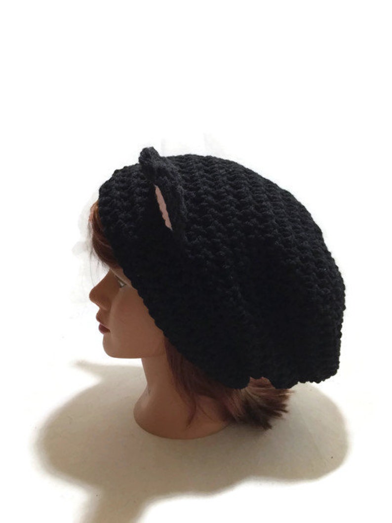 May include: Black crocheted beanie with pink cat ears. The beanie is worn on a mannequin head.