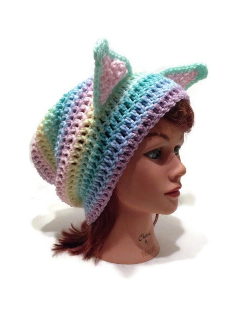 May include: A rainbow colored crochet hat with cat ears. The hat is made with a variety of pastel colors, including pink, blue, green, and yellow.