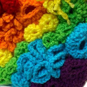 May include: A close-up of a rainbow crocheted blanket featuring a variety of stitches and textures. The blanket is made with different colours of yarn, including red, orange, yellow, green, blue, and purple.