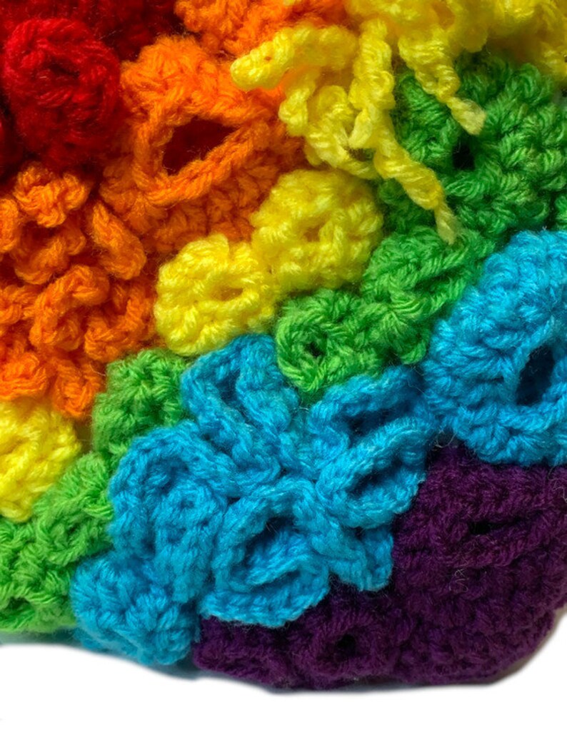 May include: A close-up of a rainbow crocheted blanket featuring a variety of stitches and textures. The blanket is made with different colours of yarn, including red, orange, yellow, green, blue, and purple.