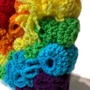 May include: A rainbow crocheted wall hanging with a textured, 3D design. The colours are red, orange, yellow, green, blue, purple, and a hint of white.