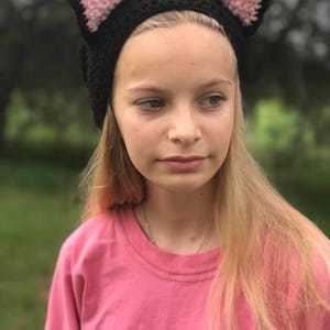 May include: A young person wearing a black crocheted cat ear headband with pink ears. The headband is worn over a pink shirt.