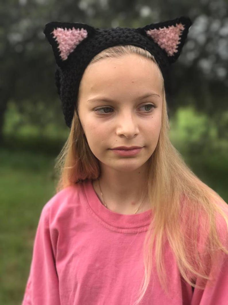 May include: A young person wearing a black crocheted cat ear headband with pink ears. The headband is worn over a pink shirt.