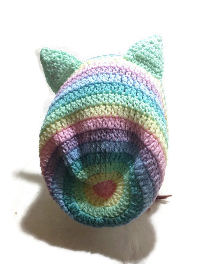 May include: A rainbow striped crocheted hat with cat ears. The hat is made with a variety of pastel colors, including pink, blue, green, and yellow.