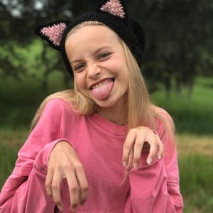 May include: A young person wearing a pink long-sleeved shirt and a black crocheted cat hat with pink ears. The person is sticking their tongue out and making a silly face.