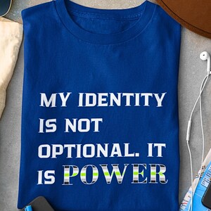 May include: A royal blue t-shirt with the text "MY IDENTITY IS NOT OPTIONAL. IT IS POWER" in white and a green, gray, and black design. The shirt is laid flat on a surface.