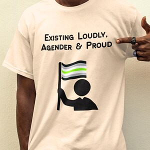 May include: Cream-colored t-shirt with the text "Existing Loudly, Agender & Proud." The shirt features a graphic of a person holding an agender pride flag. The flag has horizontal stripes of black, gray, white, and green.