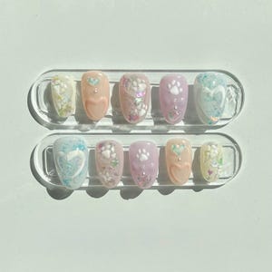 Peut inclure: Un ensemble de dix faux ongles ovales. Les ongles présentent divers motifs, notamment des cœurs, des empreintes de pattes et des motifs floraux, avec des couleurs comme le rose, le lavande et le bleu clair. Certains ongles ont des accents de paillettes.