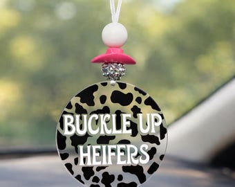 Puede incluir: Un ambientador de coche circular colgado de un espejo retrovisor, con un patrón de manchas de vaca en blanco y negro y el texto blanco BUCKLE UP HEIFERS. Está suspendido por un cordón blanco con una cuenta blanca, un disco rosa y una cuenta de estrás.