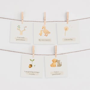 May include: Five square cards with inspirational messages are clipped to a string with wooden clothespins. The cards feature illustrations of a tree, ducklings, a dandelion, a sprout, and a puppy and kitten. Text includes phrases like "I can make good choices."