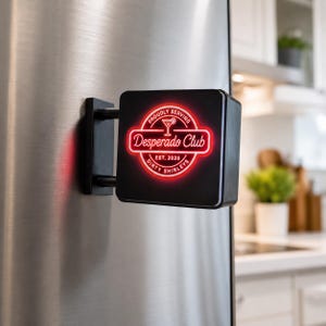 May include: A black square magnetic sign attached to a stainless steel surface. The sign features a glowing red neon-style circular logo with the text "PROUDLY SERVING", "Desperado Club", "EST. 2020", and "DIRTY SHIRLEYS" surrounding a cocktail glass icon.
