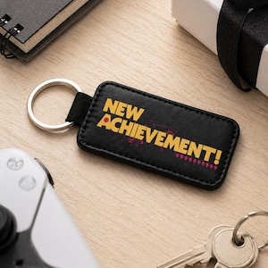 May include: Black rectangular keychain with the words "NEW ACHIEVEMENT!" in yellow and pink. The keychain has a silver key ring and a black strap. Other items on the wooden surface include a notepad, a gift box, a game controller, and keys.