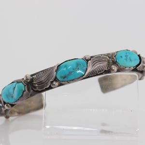 May include: Silver cuff bracelet with turquoise stones. The bracelet features oval turquoise stones set in a silver band with leaf and bead details. The open cuff design allows for easy wear.