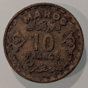 Könnte beinhalten: Eine bronzene marokkanische Münze mit dem Wort "MAROC" oben und der Zahl "10" in der Mitte. Das Wort "FRANCS" steht unten. Die Münze hat verzierte Designs an den Rändern.