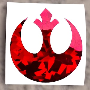 May include: A red and pink sticker featuring the Rebel Alliance symbol from Star Wars. The symbol has a textured, glittery design, set against a white background. The sticker is square-shaped.