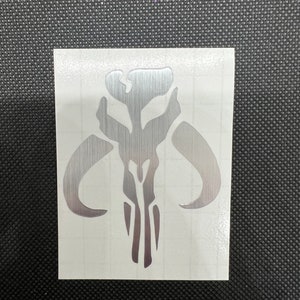 May include: Silver decal featuring the Mandalorian skull symbol. The sticker is on a white background with a grid pattern, and the design is a stylized representation of a skull with horns.
