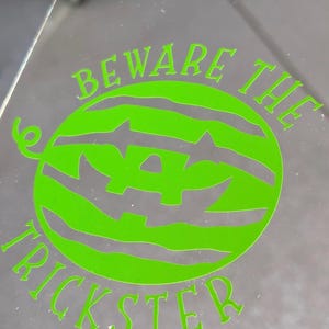 May include: A transparent surface with a neon green graphic of a watermelon carved with a jack-o'-lantern face. The text "BEWARE THE TRICKSTER" is arched around the watermelon design, perfect for Halloween.