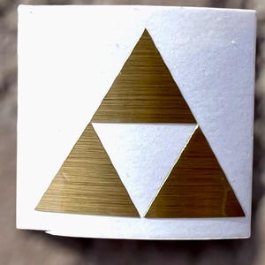 May include: A white square sticker featuring a gold-coloured Triforce symbol. The Triforce is composed of three interlocking triangles, with a textured, metallic appearance. The sticker is on a neutral background.